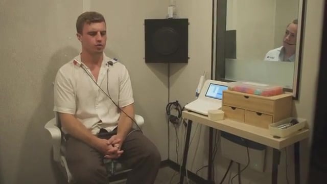 A host sitting in Fortell's soundproof hearing-test booth with sensors on his collar while an audiologist monitors from the next room